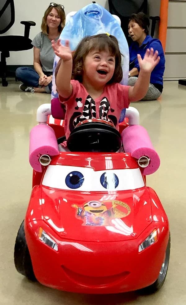 Child riding in an adapted GoBabyGo toy car with family watching