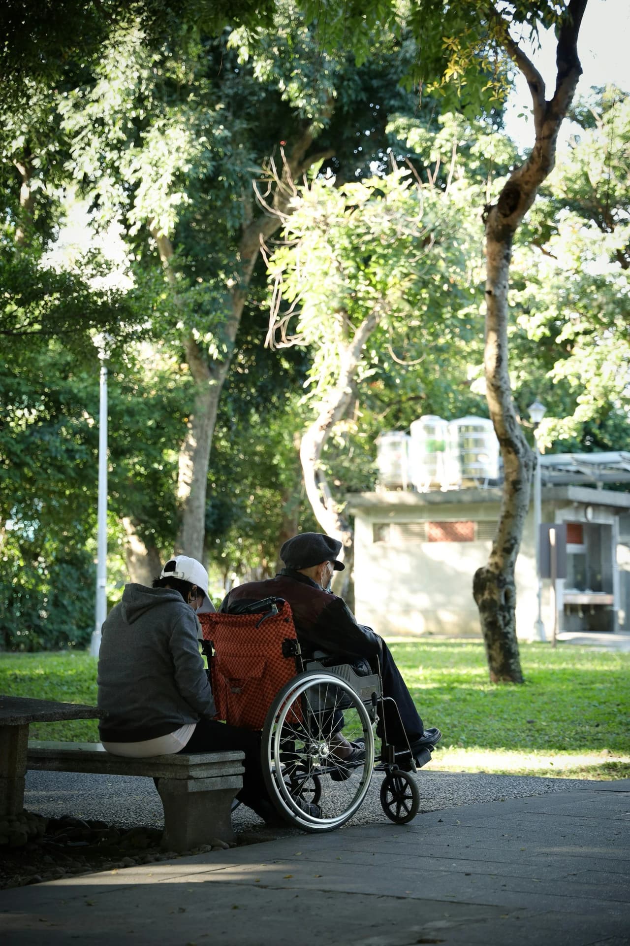 A person in a wheelchair sitting on a park bench with another person sitting nearby, surrounded by trees and sunlight filtering through the leaves.
